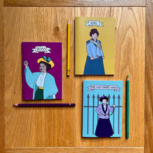 Load image into Gallery viewer, Photo of three notebooks, each next to a pencil. One notebook is purple and features a suffragette raising her first with the caption “Boss”; another is yellow with a suffragette clutching a toffee hammer with the caption “Rebel”; and a blue notebook that shows a suffragette chained to iron railings with the caption “She Who Dares, Wins”.