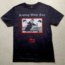 Load image into Gallery viewer, Photo of a black t-shirt featuring a black-and-white mediaeval woodcut artwork on a black background and framed in red, depicting a stereotypical crone with warts and a large nose in a black dress, holding a cane and riding a raft on water. Two black birds hover in the sky. Above it the caption reads &#39;Resting Witch Face&#39; in a white Gothic typeface. Underneath is the Clavis &amp; Claustra logo in red.