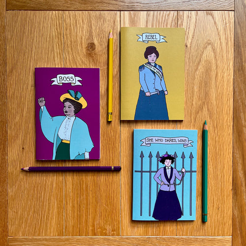 Photo of three notebooks, each next to a pencil. One notebook is purple and features a suffragette raising her first with the caption “Boss”; another is yellow with a suffragette clutching a toffee hammer with the caption “Rebel”; and a blue notebook that shows a suffragette chained to iron railings with the caption “She Who Dares, Wins”.