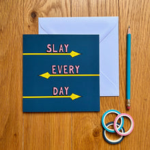 Load image into Gallery viewer, Photo of the card, which is dark blue and has the text “Slay Every Day” in pink font, each word with its own line and underlined by a yellow spear coming from alternating directions. Next to the card is a white envelope, a blue pencil, and three rolls of gift tape.