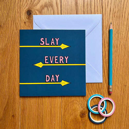 Photo of the card, which is dark blue and has the text “Slay Every Day” in pink font, each word with its own line and underlined by a yellow spear coming from alternating directions. Next to the card is a white envelope, a blue pencil, and three rolls of gift tape.