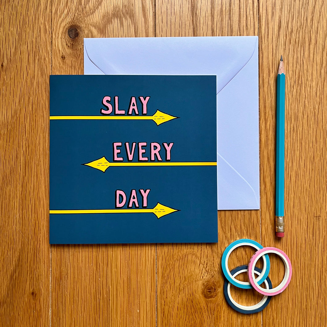 Photo of the card, which is dark blue and has the text “Slay Every Day” in pink font, each word with its own line and underlined by a yellow spear coming from alternating directions. Next to the card is a white envelope, a blue pencil, and three rolls of gift tape.