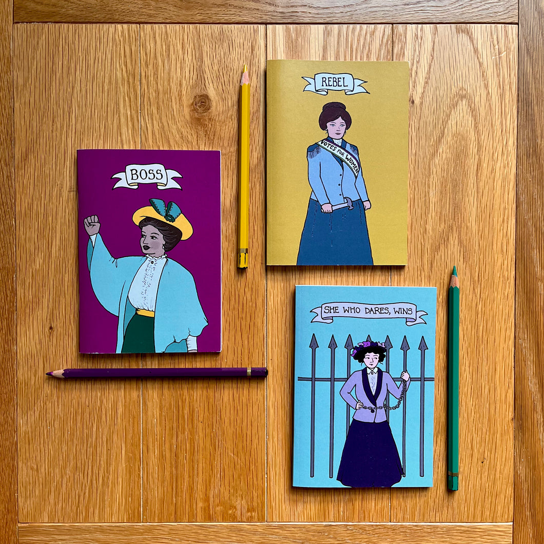 Photo of three notebooks, each next to a pencil. One notebook is purple and features a suffragette raising her first with the caption “Boss”; another is yellow with a suffragette clutching a toffee hammer with the caption “Rebel”; and a blue notebook that shows a suffragette chained to iron railings with the caption “She Who Dares, Wins”.