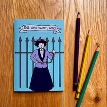 Load image into Gallery viewer, Photo of a blue notebook with shows a suffragette chained to iron railings with the caption “She Who Dares, Wins”. Three pencils are next to the notebook.
