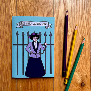 Photo of a blue notebook with shows a suffragette chained to iron railings with the caption “She Who Dares, Wins”. Three pencils are next to the notebook.