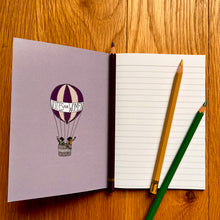 Load image into Gallery viewer, Photo of inside a notebook, which shows a blank page of lined paper with three pencils on top. The inside cover features a striped hot air balloon with a banner that says “Votes For Women” and two suffragettes waving from the basket.