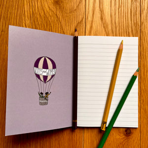 Photo of inside a notebook, which shows a blank page of lined paper with three pencils on top. The inside cover features a striped hot air balloon with a banner that says “Votes For Women” and two suffragettes waving from the basket.