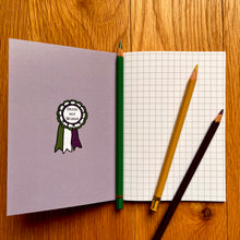 Load image into Gallery viewer, Photo of inside a notebook, which shows a blank page of squared paper with three pencils on top. The inside cover features a rosette in white, green, and purple with the text “Deeds Not Words” in the centre.