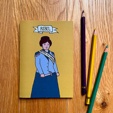 Load image into Gallery viewer, Photo of a yellow notebook with a suffragette clutching a toffee hammer with the caption “Rebel”, next to three pencils.