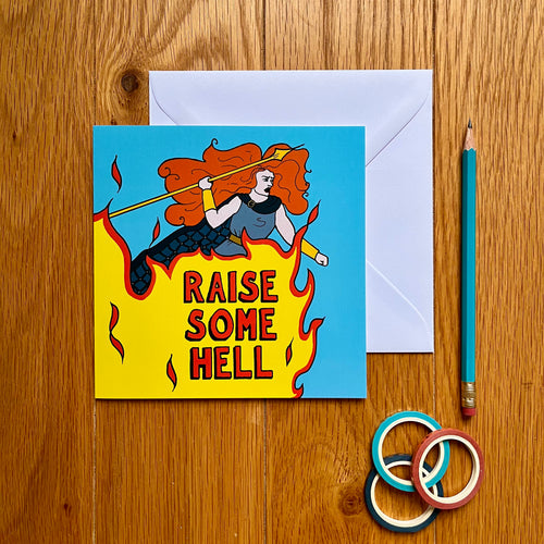 Photo of a blue card featuring Boudicca, with her ginger hair in a whirl around her as she raises a yellow spear and shouts with anger. Beneath her, a giant flame rises up with “Raise Some Hell” in red text overlaying the fire. Next to the card is a white envelope, pencil, and some rings.