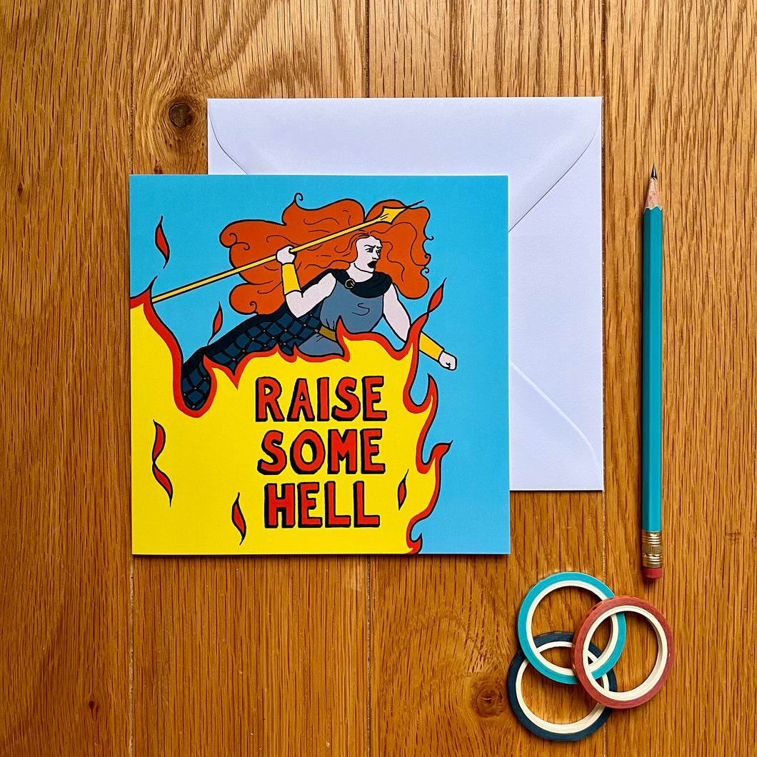 Photo of a blue card featuring Boudicca, with her ginger hair in a whirl around her as she raises a yellow spear and shouts with anger. Beneath her, a giant flame rises up with “Raise Some Hell” in red text overlaying the fire. Next to the card is a white envelope, pencil, and some rings.