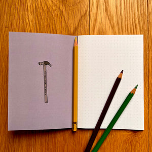 Photo of inside a notebook, which shows a blank page of dotted paper with three pencils on top. The inside cover features a toffee hammer.