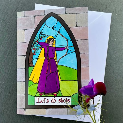 Photo of greetings card depicting a stained glass window set in a stone wall. The glass design shows a woman in purple mediaeval dress shooting a bow & arrow while birds fly above her. The caption reads 'Let's do shots'. The card is with a white envelope and two blue/purple flowers on a slate background.