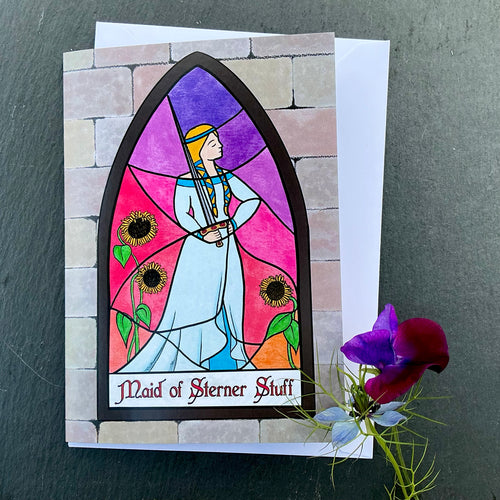Photo of greetings card depicting a stained glass window set in a stone wall. The glass design shows a woman in mediaeval dress casually wielding a sword and surrounded by sunflowers against a sunset sky. The caption reads 'Maid of Sterner Stuff'. The card is with a white envelope and two blue/purple flowers on a slate background.