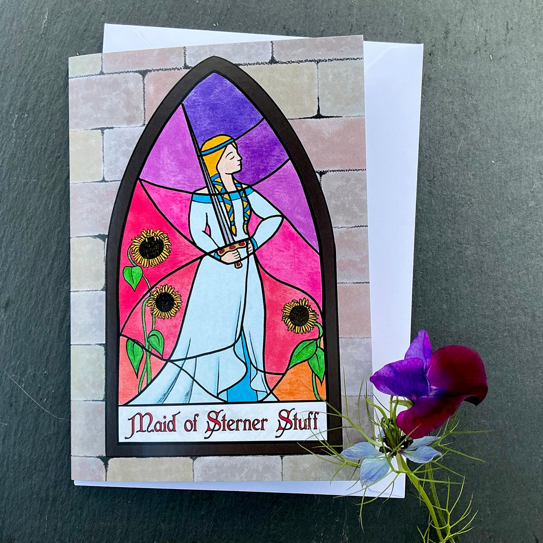 Photo of greetings card depicting a stained glass window set in a stone wall. The glass design shows a woman in mediaeval dress casually wielding a sword and surrounded by sunflowers against a sunset sky. The caption reads 'Maid of Sterner Stuff'. The card is with a white envelope and two blue/purple flowers on a slate background.