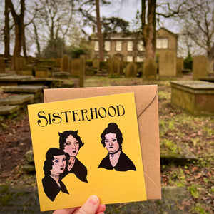 Photo of a yellow greetings card depicting the iconic portrait of the Bronte sisters but with a Goth makeover - above them is the caption "Sisterhood". Behind is the graveyard & Bronte parsonnage in Haworth.