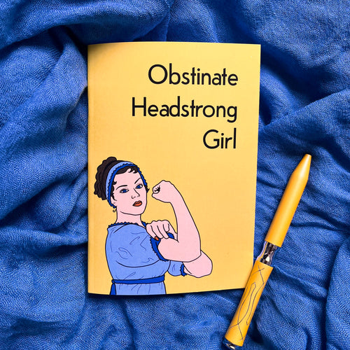 Photo of the yellow notebook which has the caption “Obstinate Headstrong Girl” next to a woman in blue Austen-era clothing doing the “Rosie the Riveter” pose of clenching her bicep. Next to the notebook is a yellow pen.