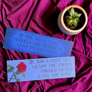 Photo of 2 bookmarks laid out next to a tiny plant. The blue bookmarks sit on a purple fabric and feature quotes from The Tenant of Wildfell Hall & Anne's poetry, with the image of a rose.