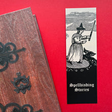 Load image into Gallery viewer, Photo of a bookmark next to a Coven Door notebook on a red background. The bookmark shows a mediaeval woodcut of a witch in a long hat standing in a circle and wielding a wand, as a demon-like creature lies on the ground. The caption beneath reads ‘Spellbinding Stories’ in a white Gothic typeface.