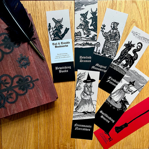 Photo of 7 bookmarks showing the 6 designs and one showing the back design, each a mediaeval woodcut with a caption. They lie on a table, next to the Coven Door notebook and a quill.