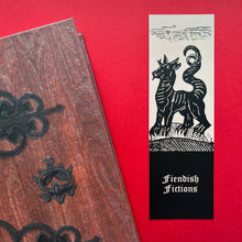 Load image into Gallery viewer, Photo of a bookmark next to a Coven Door notebook on a red background. The bookmark shows a mediaeval woodcut of a four-legged beast with a long curling tail, horns, and claws. The caption beneath reads ‘Fiendish Fictions’ in a white Gothic typeface.
