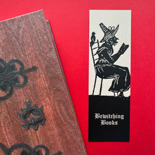 Load image into Gallery viewer, Photo of a bookmark next to a Coven Door notebook on a red background. The bookmark shows a mediaeval woodcut of a witch in a long hat sitting in a chair and reading. The caption beneath reads ‘Bewitching Books’ in a white Gothic typeface.