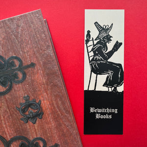 Photo of a bookmark next to a Coven Door notebook on a red background. The bookmark shows a mediaeval woodcut of a witch in a long hat sitting in a chair and reading. The caption beneath reads ‘Bewitching Books’ in a white Gothic typeface.