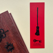 Load image into Gallery viewer, Photo of a bookmark next to a Coven Door notebook. The bookmark shows yjr back design, which is a mediaeval woodcut of a broomstick on a red background. Beneath the broomstick is the Clavis &amp; Claustra logo in black.