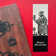 Load image into Gallery viewer, Photo of a bookmark next to a Coven Door notebook on a red background. The bookmark shows a mediaeval woodcut of a woman in a black dress and shawl contemplating a plant. A flower grows at her feet. The caption beneath reads ‘Magical Manuscripts’ in a white Gothic typeface.