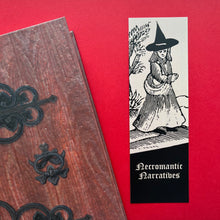 Load image into Gallery viewer, Photo of a bookmark next to a Coven Door notebook on a red background. The bookmark shows a mediaeval woodcut of a witch in a long hat standing next to a tree and smiling. The caption beneath reads ‘Necromantic Narratives’ in a white Gothic typeface.