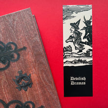 Load image into Gallery viewer, Photo of a bookmark next to a Coven Door notebook on a red background. The bookmark shows a mediaeval woodcut of a witch in a long hat sitting on a flying broomstick with the landscape below as a demon sits behind her. The caption beneath reads ‘Devilish Dramas’ in a white Gothic typeface.
