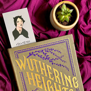 Photo of a bookmark popping out of the top of a copy of Wuthering Height, next to a tiny plant. The visible bookmark depicts Emily Bronte with a Goth makeover and "#TeamEmily" beneath.