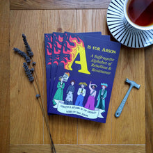 Load image into Gallery viewer, A small stack of paperback books entitled &#39;A is for Arson: A Suffragette Alphabet of Rebellion &amp; Resistance&#39; whose cover features five suffragettes marching towards the reader against a purple background, with the &#39;A&#39; of the yellow title in flames. The books sit next to a sprig of lavender, a cup of tea and a small toffee hammer.