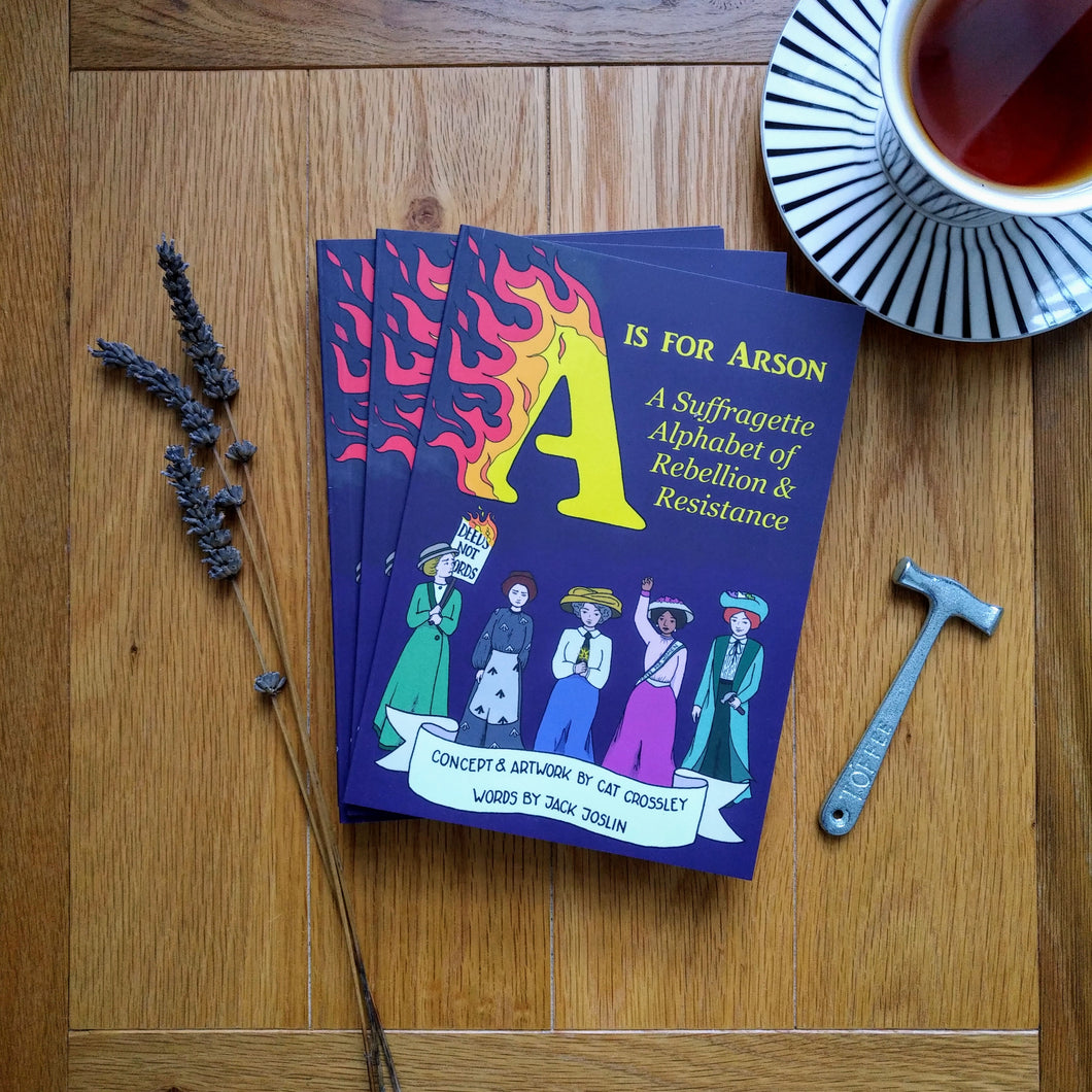 A small stack of paperback books entitled 'A is for Arson: A Suffragette Alphabet of Rebellion & Resistance' whose cover features five suffragettes marching towards the reader against a purple background, with the 'A' of the yellow title in flames. The books sit next to a sprig of lavender, a cup of tea and a small toffee hammer.