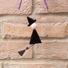 Load image into Gallery viewer, Photo of glasswork which depicts a witch in a minimalist style, with a black dress and black hat, flying on a broomstick. The artwork hands from a purple thread against a brick wall.