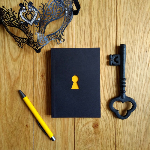 Photograph of the Escape Journal paperback book with a black cover and yellow keyhole cutout, pictured with a yellow pen, masquerade mask, and iron key.