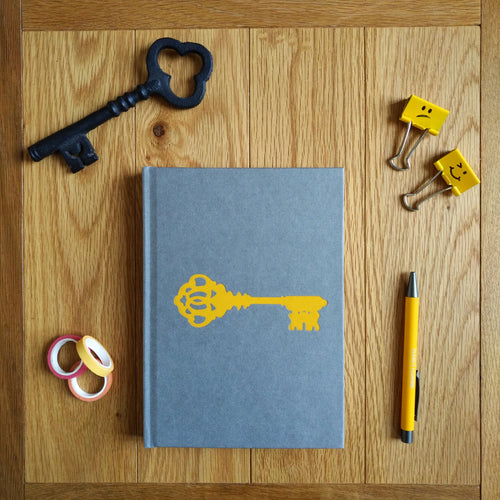 Photo of the journal, a grey hardback with a yellow ornate key on the cover. Next to the journal is a black metal key, some gift tape, a yellow pen, and some yellow document clips.