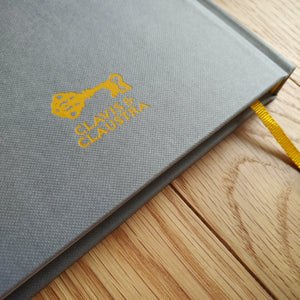 Photo of the journal, a grey hardback with the yellow Clavis & Claustra key logo at the base. A yellow page saver ribbon emerges from the pages.