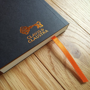 Photograph of the back of the black Escape Journal paperback, showing the Clavis & Claustra key logo in gold foil, with an orange ribbon placeholder emerging from the pages.