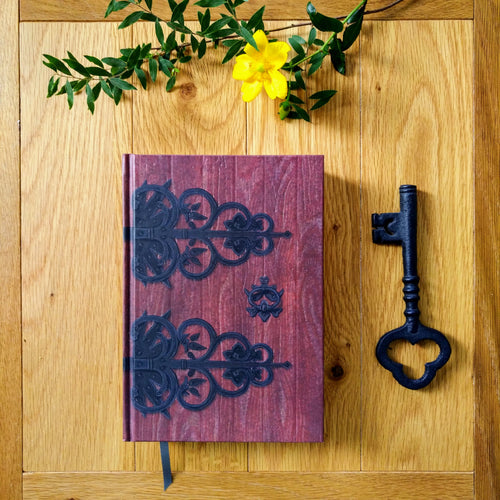 Photo of a hardback notebook styled as a deep read wooden door with ornate black metal decorations and a keyhole. Next to the book is a black metal key and some flowers.