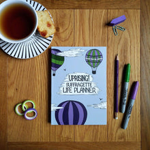 Load image into Gallery viewer, Photo of the light blue planner, which shows three hot air balloons in suffragette colours in the sky with the title “Uprising! Suffragette Life Planner” as text within a cloud. Next to the notebook are three pens, some gift tape, some sticky labels, paper clips, and a cup of black tea on a saucer with a slice of cake.