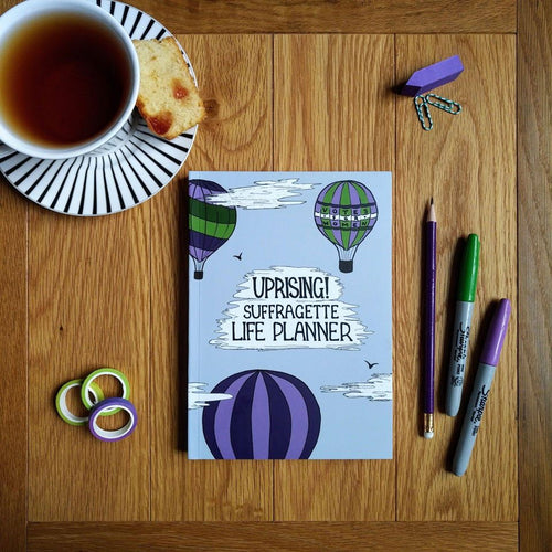 Photo of the light blue planner, which shows three hot air balloons in suffragette colours in the sky with the title “Uprising! Suffragette Life Planner” as text within a cloud. Next to the notebook are three pens, some gift tape, some sticky labels, paper clips, and a cup of black tea on a saucer with a slice of cake.
