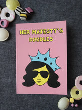 Load image into Gallery viewer, Photo of Her Majesty’s Doodles notebook, featuring a pink cover with the title in yellow and a stylised illustration of an unsmiling woman with long black hair, wearing sunglasses and an extravagant blue crown. Next to the notebook are an assortment of sweets and biscuits.