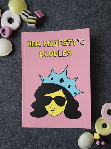 Photo of Her Majesty’s Doodles notebook, featuring a pink cover with the title in yellow and a stylised illustration of an unsmiling woman with long black hair, wearing sunglasses and an extravagant blue crown. Next to the notebook are an assortment of sweets and biscuits.