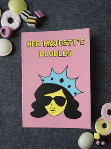 Photo of Her Majesty’s Doodles notebook, featuring a pink cover with the title in yellow and a stylised illustration of an unsmiling woman with long black hair, wearing sunglasses and an extravagant blue crown. Next to the notebook are an assortment of sweets and biscuits.
