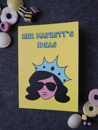 Photo of Her Majesty’s Ideas notebook, featuring a yellow cover with the title in blue and a stylised illustration of an unsmiling woman with long black hair, wearing sunglasses and an extravagant blue crown. Next to the notebook are an assortment of sweets and biscuits.