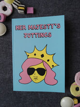 Load image into Gallery viewer, Photo of Her Majesty’s Jottings notebook with the title in pink, featuring a blue cover with a stylised illustration of an unsmiling woman with long pink hair, wearing sunglasses and an extravagant yellow crown. Next to the notebook are an assortment of sweets and biscuits.