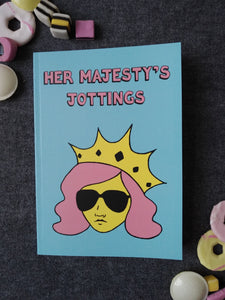 Photo of Her Majesty’s Jottings notebook with the title in pink, featuring a blue cover with a stylised illustration of an unsmiling woman with long pink hair, wearing sunglasses and an extravagant yellow crown. Next to the notebook are an assortment of sweets and biscuits.