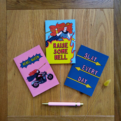 Three notebooks. The Rebel Queen pink notebook shows Boudicca holding a spear and riding a motorcycle; the Raise Some Hell light blue notebook has Boudicca holding a spear as a fire erupts beneath her; and the dark blue Slay Every Day has that as a caption underlined by yellow spears for each word. Next to the notebooks is a pink pen.