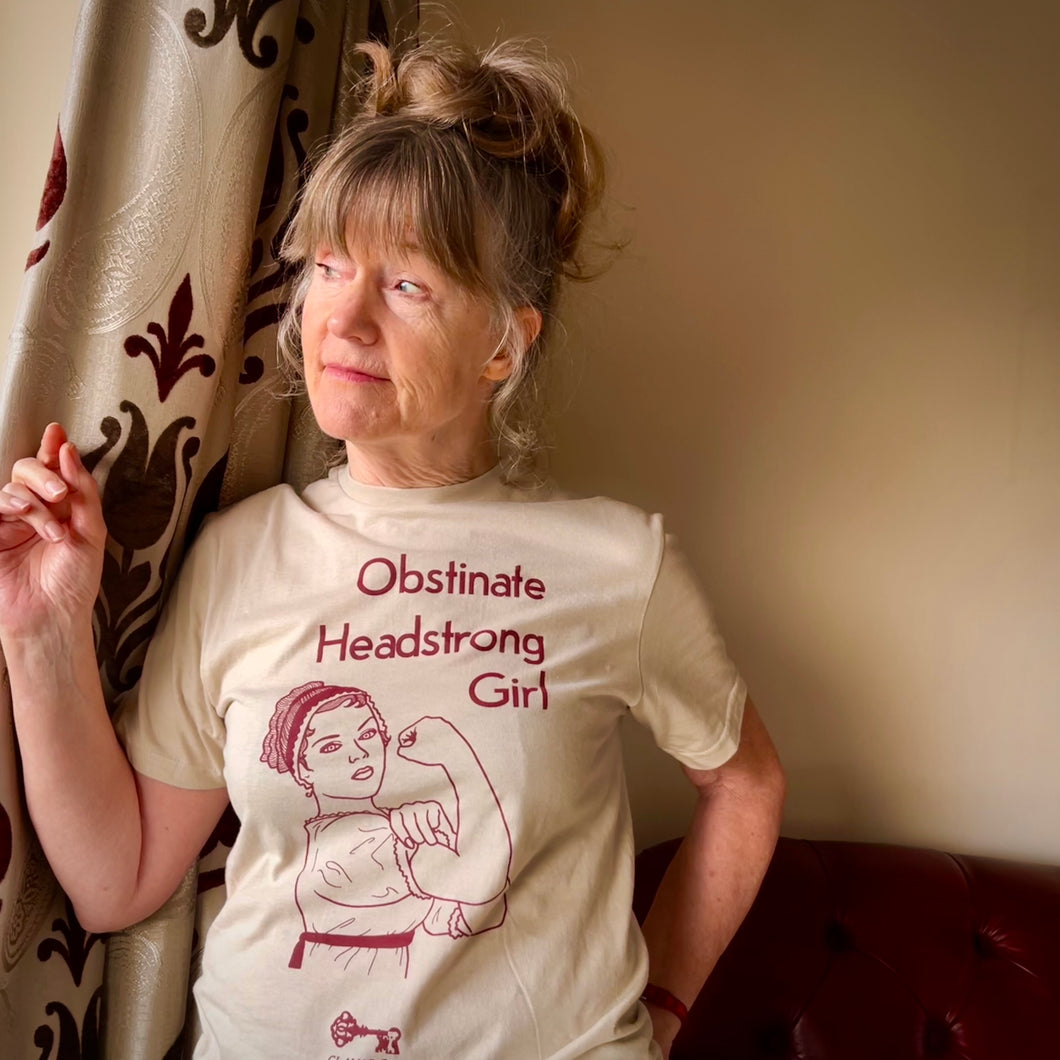 Photograph of a t-shirt modelled by a white-skinned woman with long silvery brown hair as she looks out the window. The cream coloured t-shirt features a drawing in red depicting a woman in Jane Austen-style clothing in the “Rosie the Riveter” pose, curling an arm and clutching her bicep triumphantly. Above in rest text are the words 